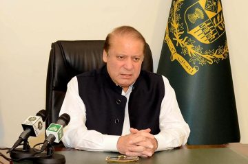 A man seated at a desk, wearing a white shalwar kameez and dark waistcoat, speaking in front of microphones with the official Government of Pakistan emblem displayed behind him.
