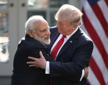 Two senior political figures stand outdoors, embracing each other in a warm, formal hug. One man has white hair and a white beard, wearing a dark suit, while the other man has light blond hair and is also dressed in a dark suit with a red tie. A blurred American flag is visible in the background.