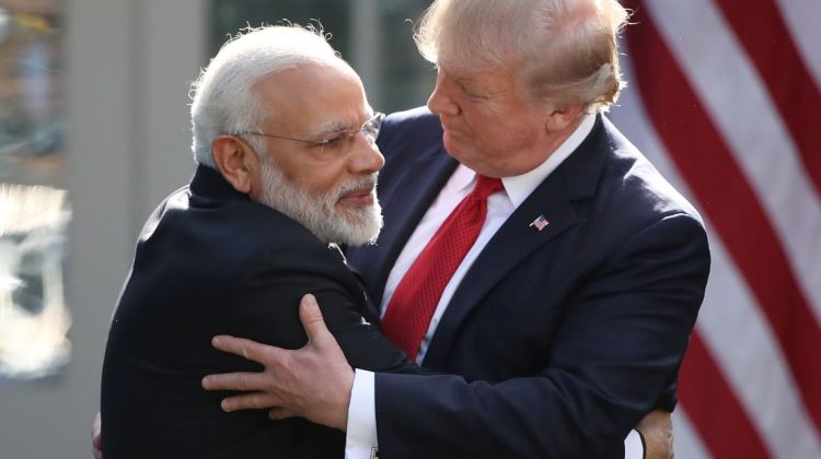 Two senior political figures stand outdoors, embracing each other in a warm, formal hug. One man has white hair and a white beard, wearing a dark suit, while the other man has light blond hair and is also dressed in a dark suit with a red tie. A blurred American flag is visible in the background.