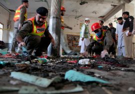 Rescue workers and police officers examine debris inside a damaged building after a violent attack, searching for evidence among burnt and scattered materials.