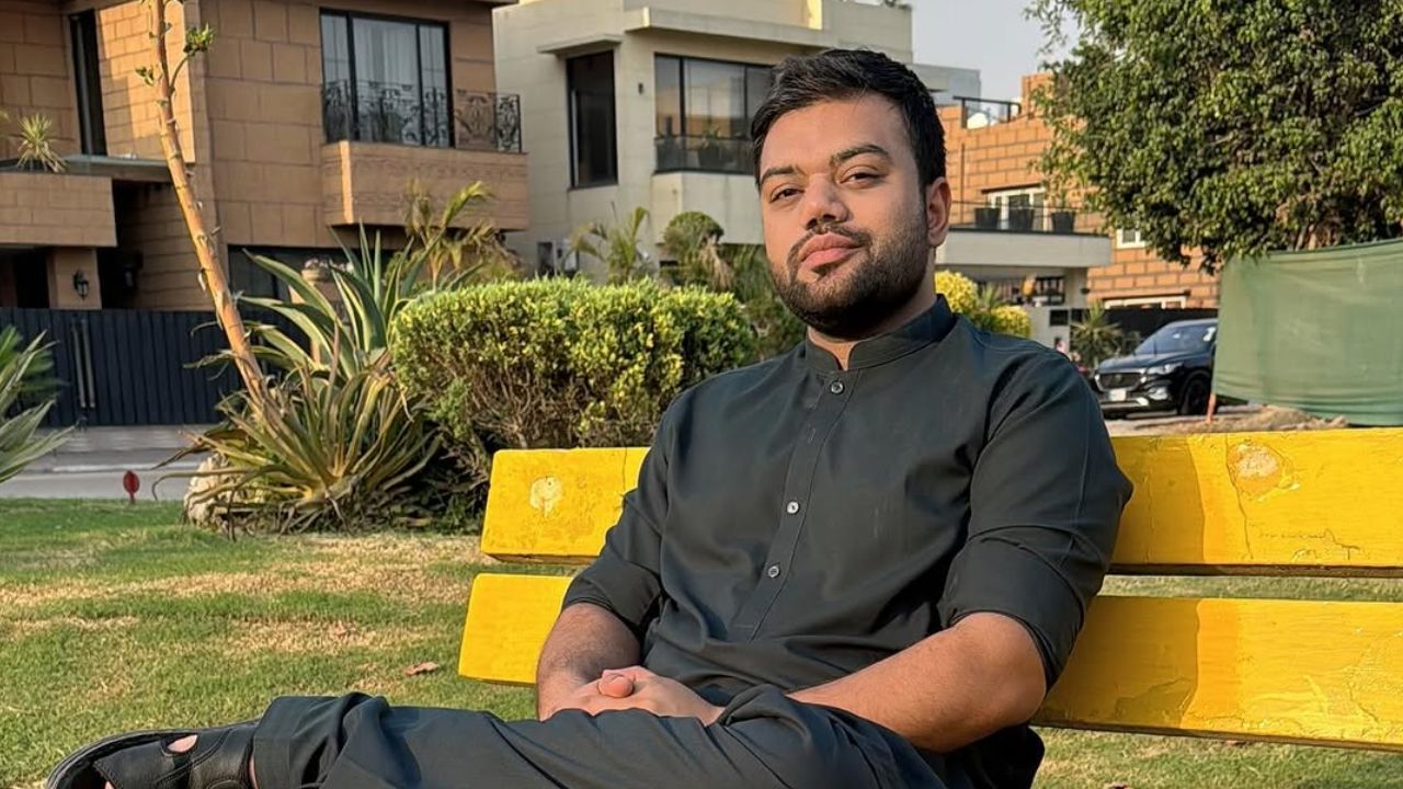 A man wearing a black shalwar kameez sits confidently on a yellow park bench, with modern houses, plants, and greenery visible in the background during daylight.