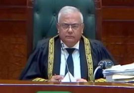A senior Pakistani judge wearing official judicial robes sits at the bench in a courtroom, looking forward with documents and files placed beside him.