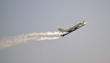 A Tejas fighter jet climbs sharply into the sky during an aerial maneuver, leaving a white smoke trail behind it. The aircraft is seen against a clear blue sky, showcasing its performance capabilities.