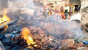 A destroyed chemical factory site in Faisalabad with thick smoke and flames rising from scattered debris. Rescue workers in safety gear stand among collapsed walls and rubble while searching for victims.