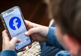 A young person holding a smartphone with the Facebook app open on the screen while typing.