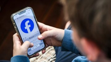A young person holding a smartphone with the Facebook app open on the screen while typing.