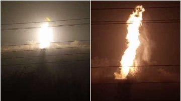A large, intense column of fire rising vertically into the night sky, captured from a distance with power lines visible in the foreground. Heavy smoke surrounds the bright flames.