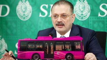 A government official speaking at a press briefing with an image of a pink public transport bus placed in front.