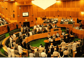 A view of the Azad Jammu and Kashmir Legislative Assembly during a session, with members standing and engaging in discussion inside the large wooden-paneled hall.