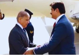 A welcoming moment at the airport showing two officials shaking hands as one arrives; a security personnel holds an umbrella in the background near an aircraft