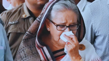 An elderly woman wearing glasses and a patterned shawl wipes her nose with a tissue while appearing emotional, surrounded by people in the background.