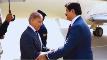 A welcoming moment at the airport showing two officials shaking hands as one arrives; a security personnel holds an umbrella in the background near an aircraft