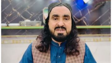 A man with long black hair, a full beard, and a traditional Sindhi cap is sitting indoors, looking directly at the camera. He is wearing a blue kurta and a brown checkered waistcoat, with a marble-patterned wall behind him.