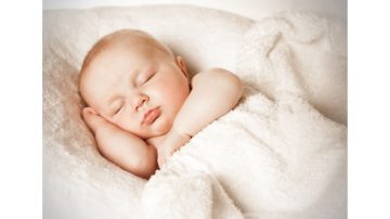 A peacefully sleeping baby wrapped in a soft white blanket, resting with one hand under the cheek.