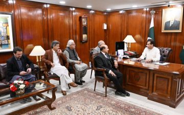 A formal meeting inside a wood-paneled government office where five men are seated. One man sits behind a large wooden desk, appearing to lead the discussion. The others sit on chairs and a sofa in front of him. A Pakistani flag and a portrait of a national leader hang on the wall. Lamps, files, and office decor are visible around the room.