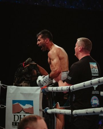 A victorious boxer stands inside the ring, shirtless and wearing white gloves, shouting in celebration while a coach or corner man stands beside him. A cameraman is visible in the background capturing the moment. The scene is lit dramatically with the crowd in darkness.