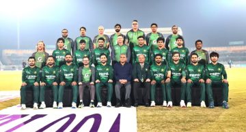 A group photo of the Pakistan national cricket team and staff, all dressed in green team kits, posing together on a cricket field at night. The players and officials are arranged in three rows, smiling towards the camera under stadium lights.