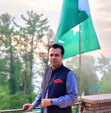 “A man standing on a balcony wearing a traditional dark waistcoat over a light blue kurta, with a Pakistan flag waving behind him and tall green trees in the background.”