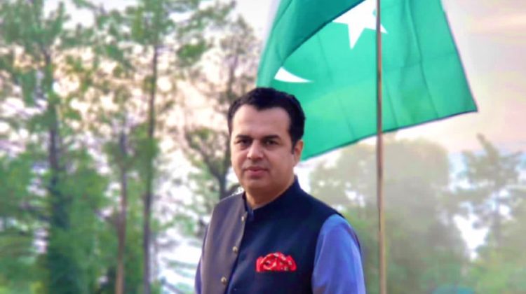 “A man standing on a balcony wearing a traditional dark waistcoat over a light blue kurta, with a Pakistan flag waving behind him and tall green trees in the background.”