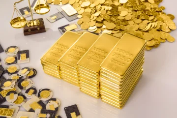 "Stacks of shiny gold bars arranged in rows with gold coins, small minted gold pieces, and a weighing scale placed around them on a reflective surface."