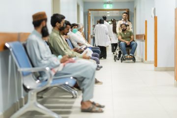 "Patients seated in a hospital waiting area while a man in a wheelchair is pushed by an attendant down the hallway, with doctors and staff visible in the background."