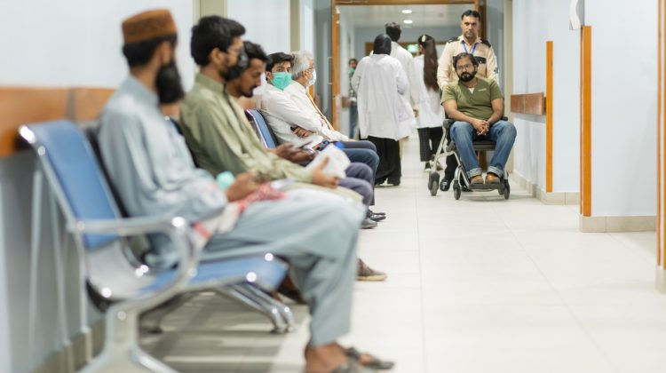 "Patients seated in a hospital waiting area while a man in a wheelchair is pushed by an attendant down the hallway, with doctors and staff visible in the background."