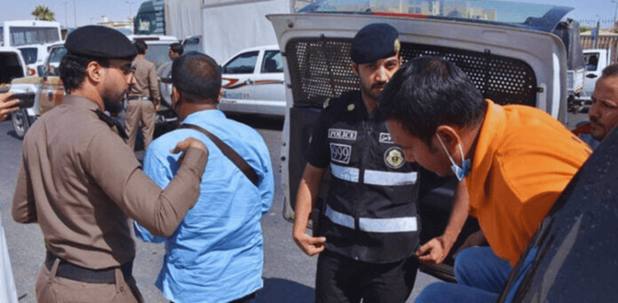 Saudi police officers detain and escort several individuals during a crackdown on illegal expatriates, with detainees being moved toward a police vehicle.