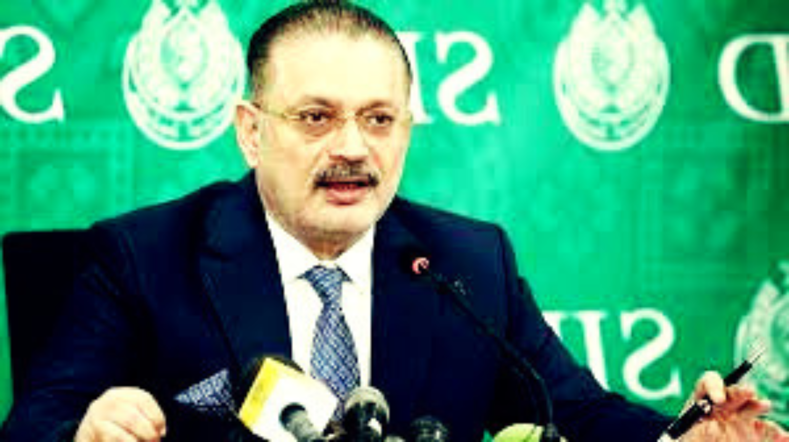 Sindh Minister Sharjeel Inam Memon addressing a press conference, speaking into microphones with the Sindh government emblem visible on the green backdrop behind him.