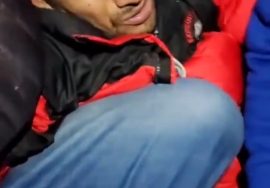 A distressed man in a red and black jacket crouched tightly in a corner, his face showing fear and pain as others stand close around him.