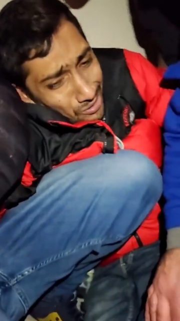 A distressed man in a red and black jacket crouched tightly in a corner, his face showing fear and pain as others stand close around him.