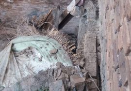 Debris of a collapsed mud-and-wood roof is shown on the ground, with broken bricks, wooden beams, straw, and roofing material scattered inside the damaged structure.