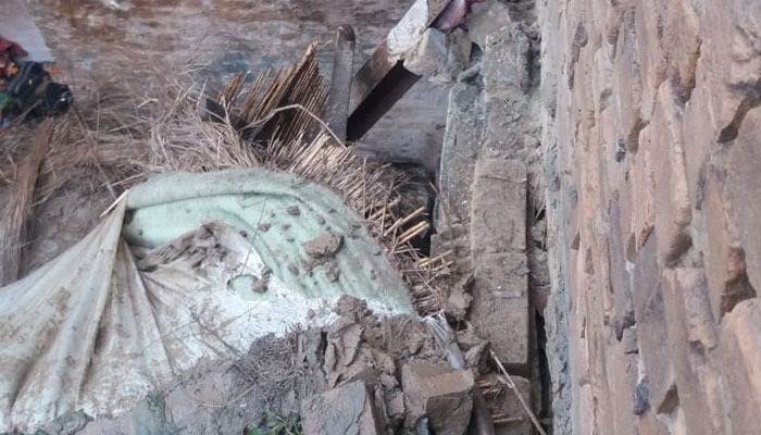 Debris of a collapsed mud-and-wood roof is shown on the ground, with broken bricks, wooden beams, straw, and roofing material scattered inside the damaged structure.