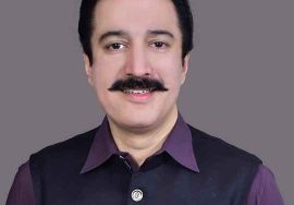 A portrait of a man wearing a dark waistcoat over a purple shirt, smiling at the camera against a plain grey background.