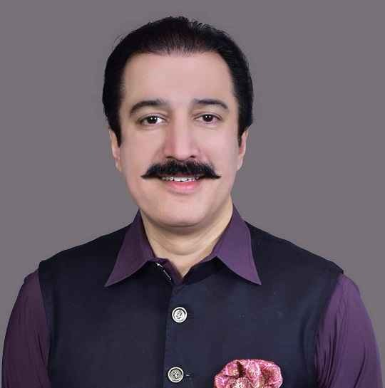 A portrait of a man wearing a dark waistcoat over a purple shirt, smiling at the camera against a plain grey background.