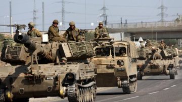 A convoy of armored military vehicles and tanks moving along a highway, with several soldiers in uniform positioned on top and inside the vehicles.