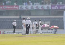 Cricketers and umpires stand in the middle of the field during the Bangladesh vs Ireland Test match as play is halted due to earthquake tremors. The stadium seats in the background appear partially empty, and sunlight falls across the ground.