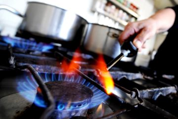 A close-up shot of a gas stove burner with blue flames, while a person ignites another burner using a lighter. A cooking pot is placed on the stove in the background.