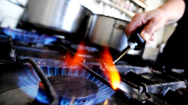 A close-up shot of a gas stove burner with blue flames, while a person ignites another burner using a lighter. A cooking pot is placed on the stove in the background.