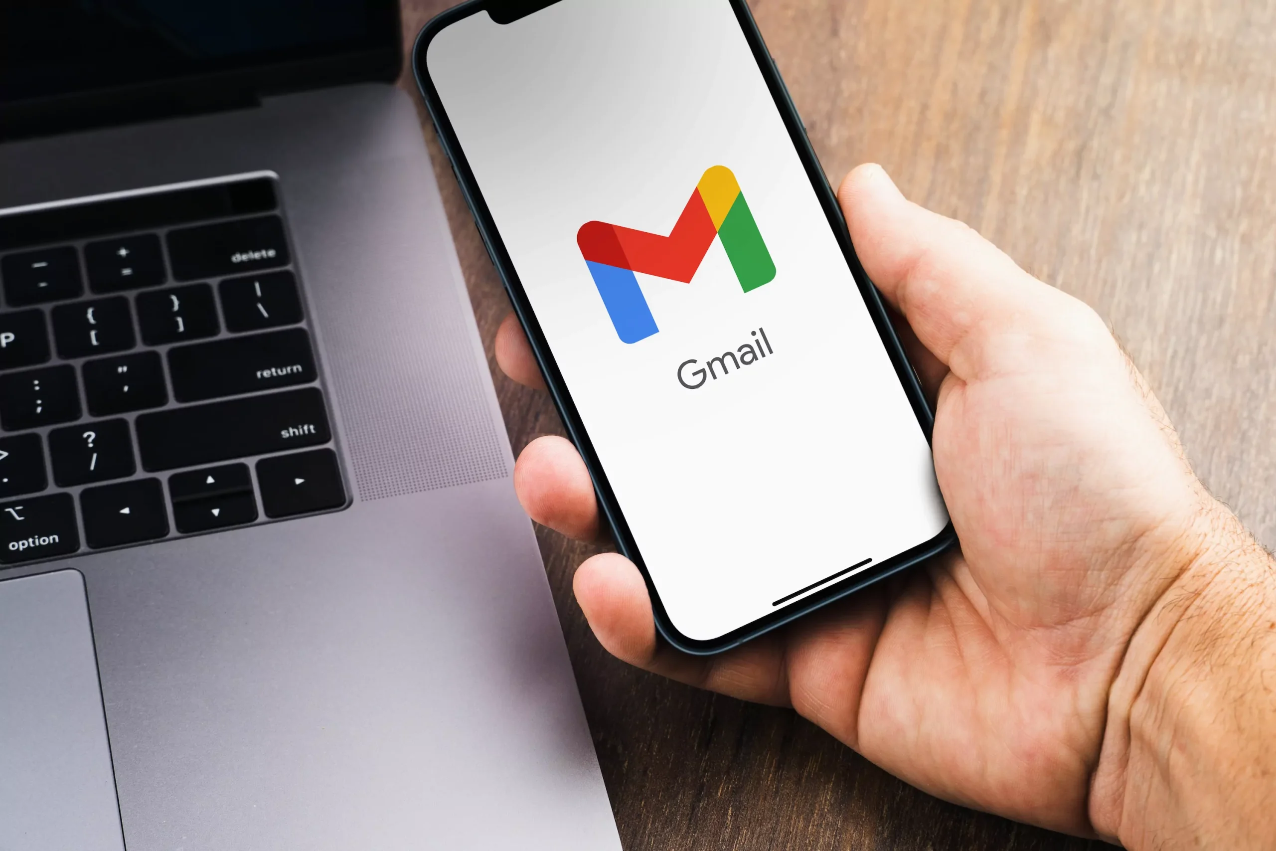 A hand holding a smartphone displaying the Gmail logo on the screen, placed next to an open laptop keyboard on a wooden desk.