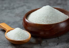 A wooden bowl filled with granulated white sugar placed on a textured gray surface, with a wooden spoon holding additional sugar next to it.