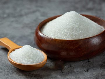 A wooden bowl filled with granulated white sugar placed on a textured gray surface, with a wooden spoon holding additional sugar next to it.