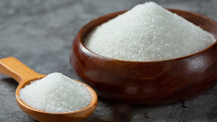 A wooden bowl filled with granulated white sugar placed on a textured gray surface, with a wooden spoon holding additional sugar next to it.