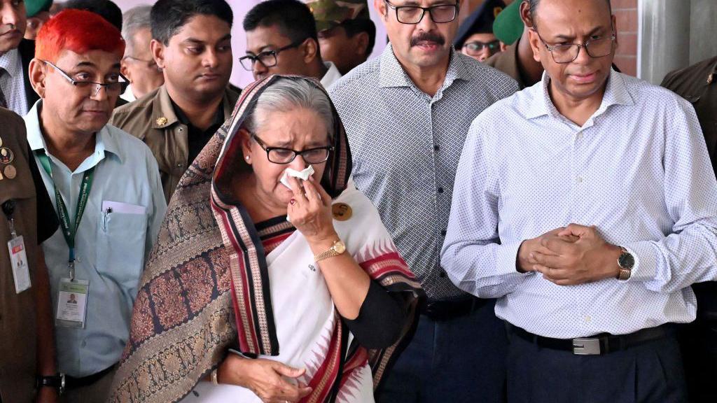 A senior woman wearing a traditional South Asian saree wiping her face while surrounded by security personnel and officials.