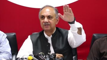 “Press conference mein baithay aik Pakistani siyasi shaksiyat jis ka haath utha hua hai, samne microphones lage hue hain, red background ke sath.”