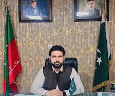 A Pakistani official seated at a desk with the Pakistan flag and a party flag displayed behind him. Portraits of Quaid-e-Azam Muhammad Ali Jinnah and another leader hang on the wall above.