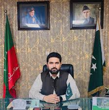 A Pakistani official seated at a desk with the Pakistan flag and a party flag displayed behind him. Portraits of Quaid-e-Azam Muhammad Ali Jinnah and another leader hang on the wall above.