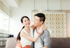 A smiling Asian couple sitting closely on a sofa, with the woman embracing the man from the side. Both appear happy and relaxed in a bright, modern living room.