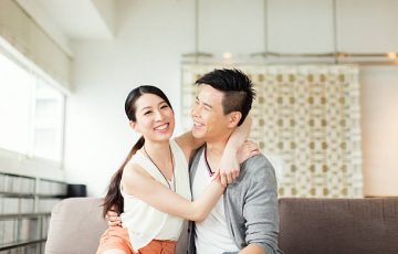 A smiling Asian couple sitting closely on a sofa, with the woman embracing the man from the side. Both appear happy and relaxed in a bright, modern living room.