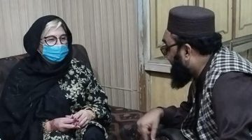 A woman wearing a black headscarf, floral dress, and a blue surgical mask sits on a sofa while speaking to a bearded man in traditional attire inside a modest room.