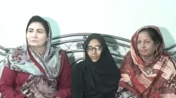 A young girl sitting between two women on a bed, all wearing traditional South Asian clothing, posing calmly indoors.